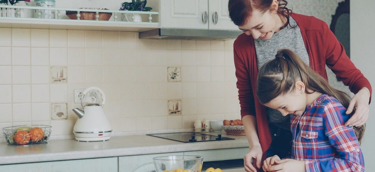 Young pretty mother teaching her cute daughter to cut vegetables properly. Little smiling girl cooking together with loving mom at home in modern kithcen