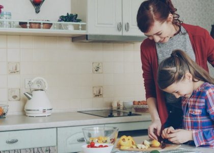 Young pretty mother teaching her cute daughter to cut vegetables properly. Little smiling girl cooking together with loving mom at home in modern kithcen